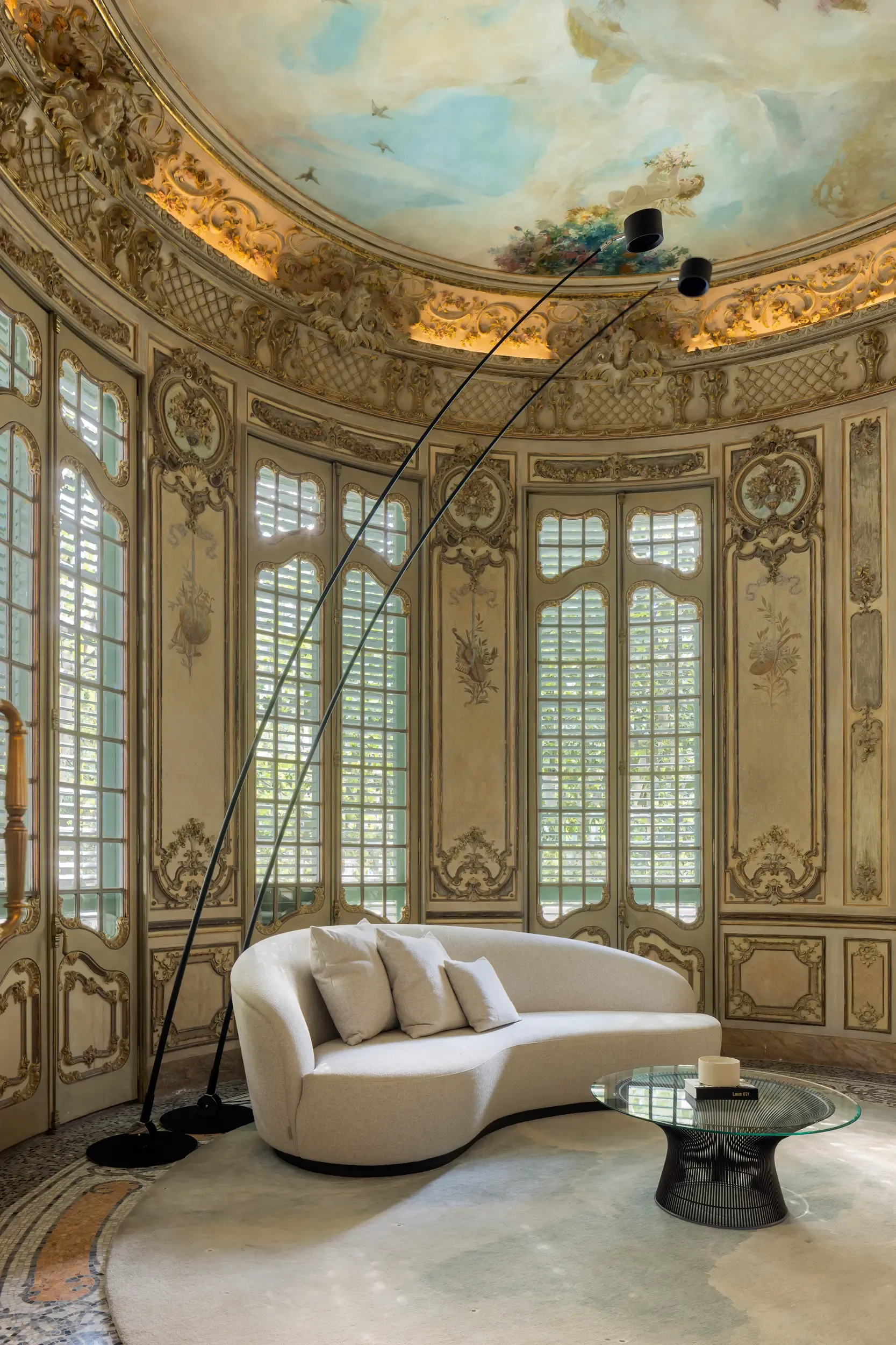 Elegant lounge with curved cream sofa, glass coffee table, and ornate painted ceiling surrounded by detailed Art Nouveau wall panels and tall windows.