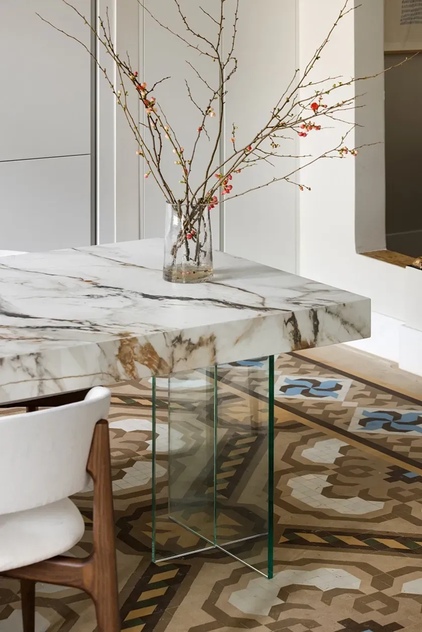 Marble table with glass legs and a vase with branches on patterned tiled floor.