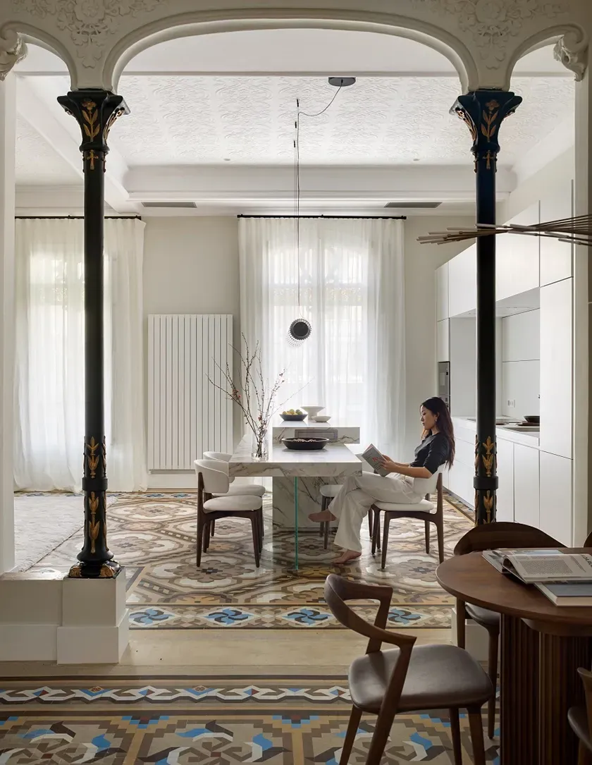 Woman reading at the marble dining island in a refined kitchen space with Art Nouveau columns and mosaic flooring.