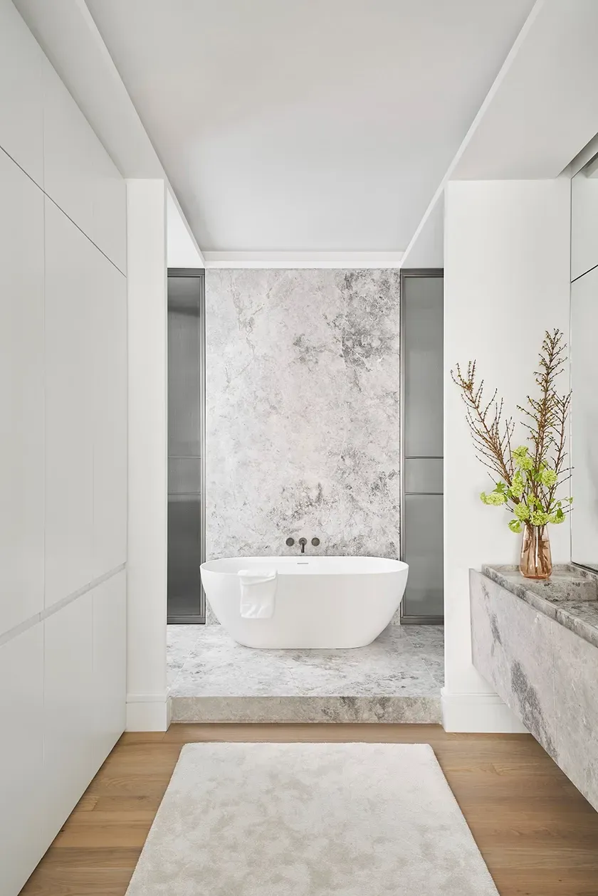 Minimalist bathroom with white freestanding tub, marble textures, soft lighting, and delicate decorative branches in a copper-toned vase.
