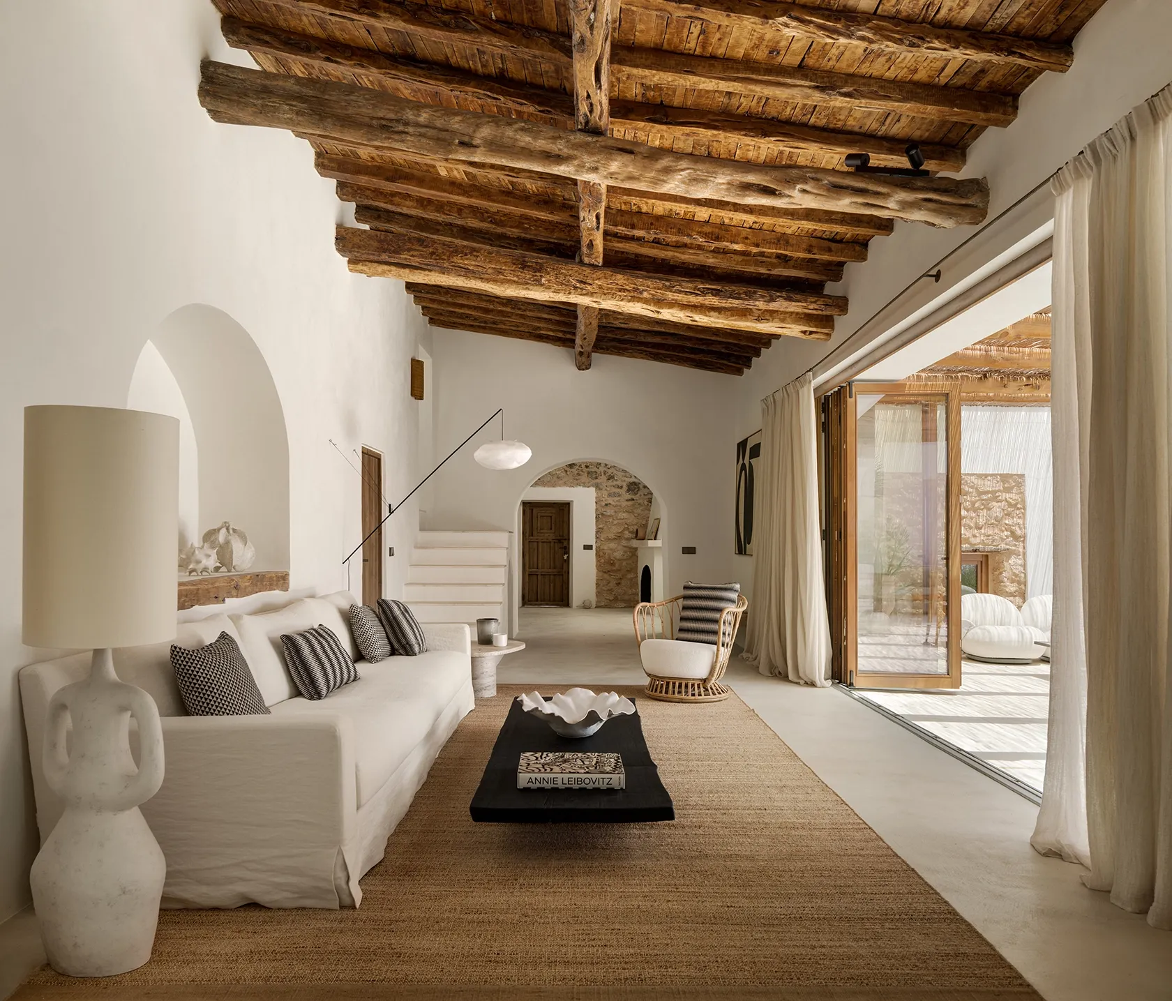 Spacious living room with natural light, raw wooden beams, minimalist furniture, and smooth connection to the outdoor lounge area.