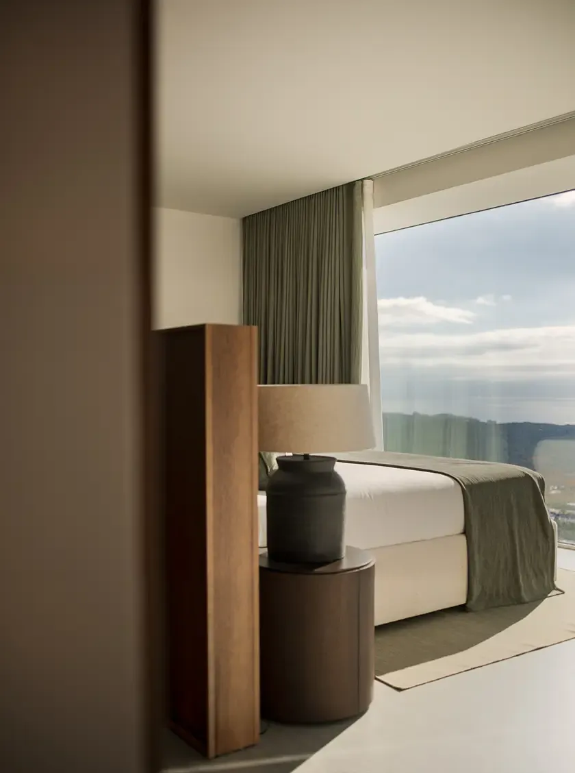 A luxurious bedroom with a peaceful ambiance, offering expansive views through a large window. The minimalist decor with earthy tones and natural textiles creates a calm, inviting atmosphere.
