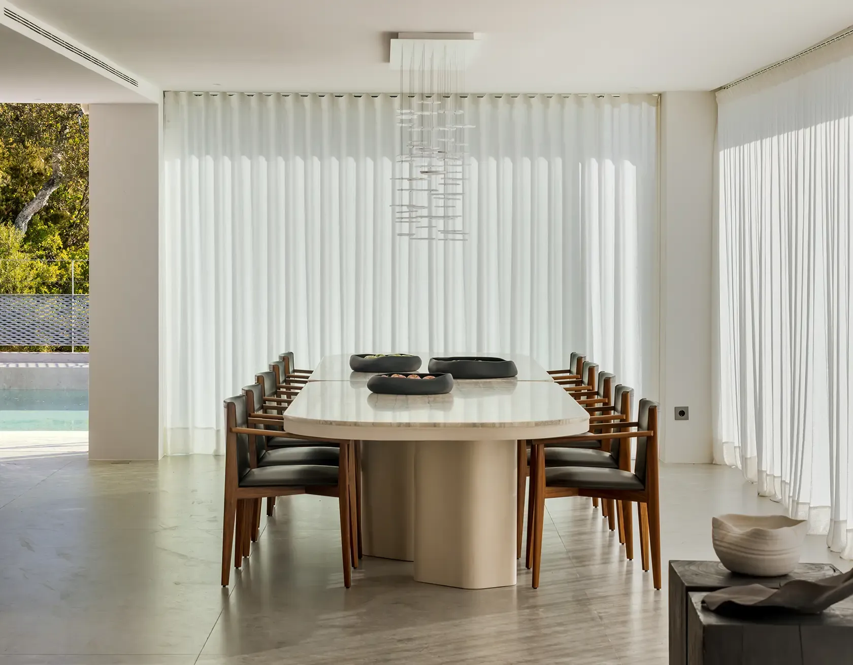 Modern dining room with a long marble table, minimalist chairs, and sculptural lighting, perfectly framed by expansive windows offering breathtaking views of the hills and sky beyond.