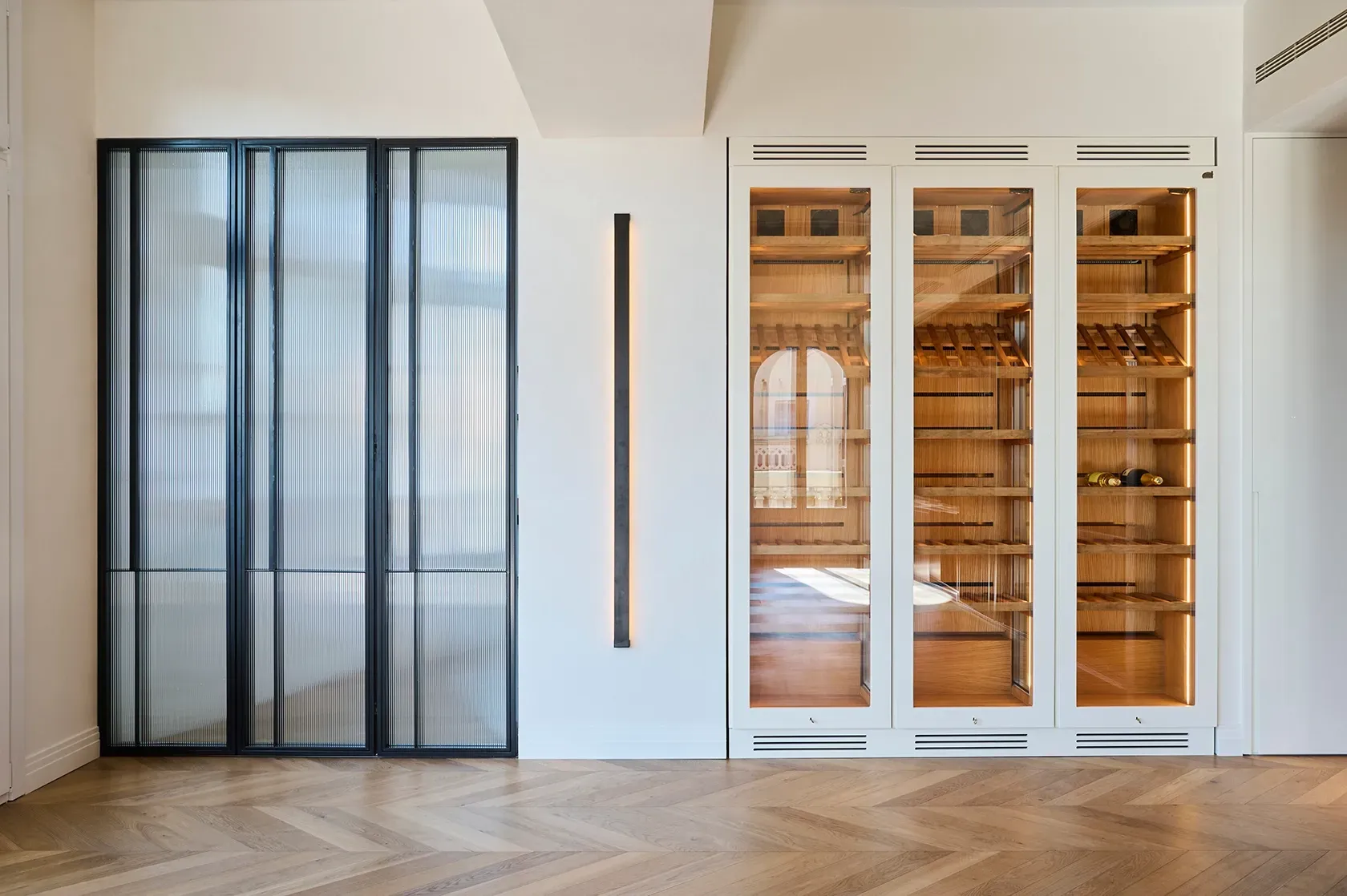 Custom wine cellar with illuminated wood shelving and glass doors, next to industrial-style black-framed panels on herringbone oak flooring.